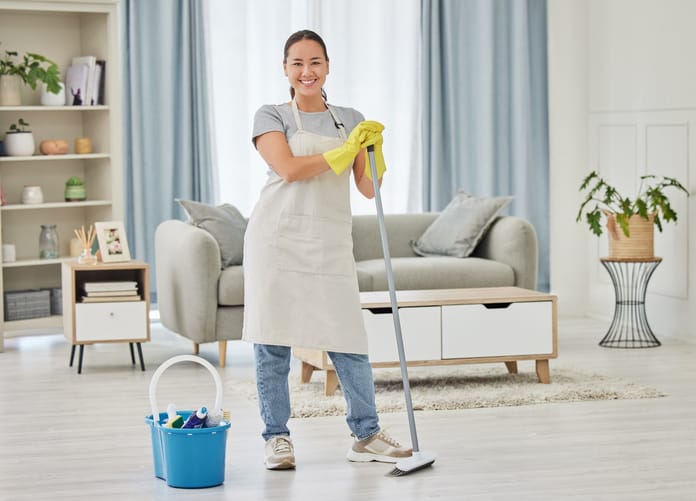 A happy smiling mixed race woman cleaning the floor of her apartment. One Asian woman using a broom and a bucket of cleaning supplies to clean the floor
